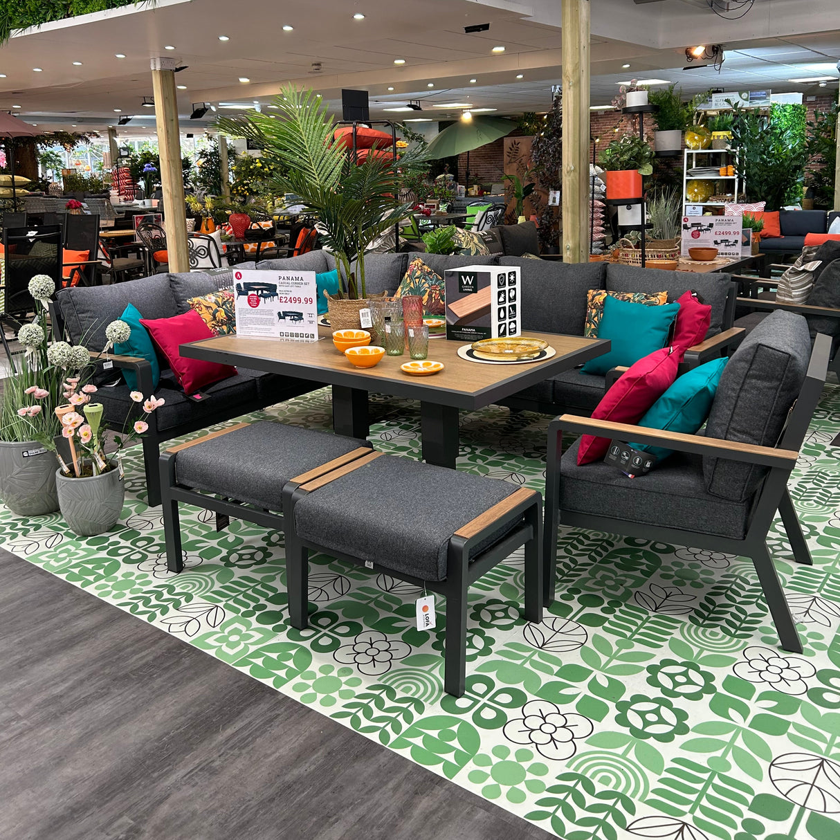 A modern Panama modular rectangular casual corner set displayed in an showroom setting, consisting of a black frame with colorful cushions, a wood effect ceramic glass tabletop, and two stools with cushions.